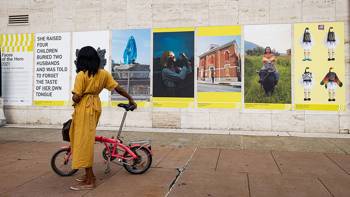 A woman holding a bicycle and walking on the sidewalk in front of a wall of artistic posters